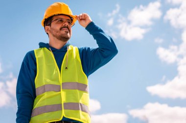 Sarı şapkalı, gözlüklü, yeşil yelekli, eli kasklı, uzaklara bakan Latin bir işçi.