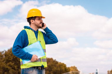 Sarı şapkalı, gözlüklü, yeşil yelekli, telefonda konuşan ve elinde bir klasör tutan Latin bir işçi.