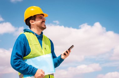 Sarı şapkalı, gözlüklü, yeşil yelekli, elinde bir klasörle telefonla konuşan ve gülümseyen Latin bir işçi..