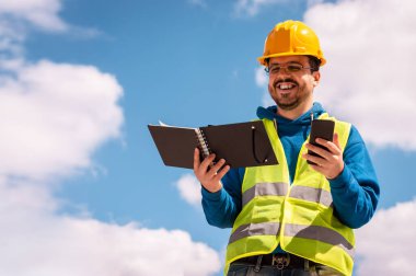 Sarı şapkalı, gözlüklü, yeşil yelekli, kitap okuyan, telefon eden ve gülümseyen Latin bir işçi..