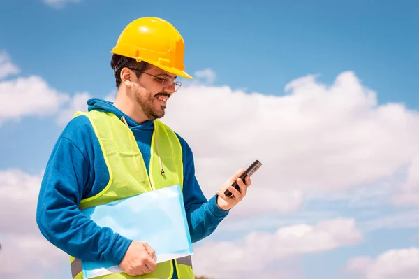 Latin worker with yellow hard hat, glasses and green vest, talking on the phone and holding a folder in his hand