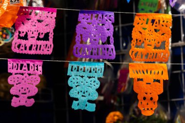 Üzerinde insanların isimlerinin yazılı olduğu renkli kağıt bayraklar. Bayraklar kabloda asılı. Meksika Ölüler Günü geleneği, adaklar, yemek, renkler, dekorasyonlar ve papel picado, La Catrina.