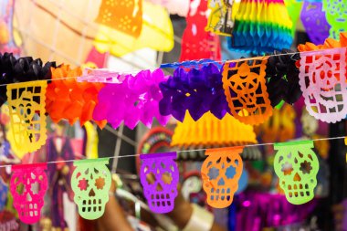 Renkli bir kağıt kafatası ve afiş gösterisi. Görüntü renk ve şekillerin bir karışımıdır. Meksika Ölüler Günü geleneği, adaklar, yemek, renkler, dekorasyonlar ve papel picado, La Catrina.