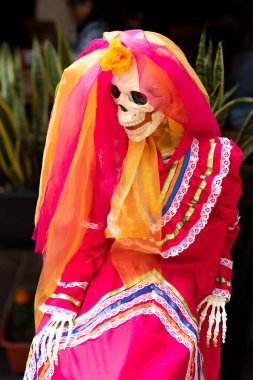 Kırmızı elbiseli, duvağı ve çiçekleri olan bir iskelet. İskelet bir sandalyede oturuyor. Meksika Ölüler Günü geleneği, adaklar, yemek, renkler, dekorasyonlar ve papel picado, La Catrina.