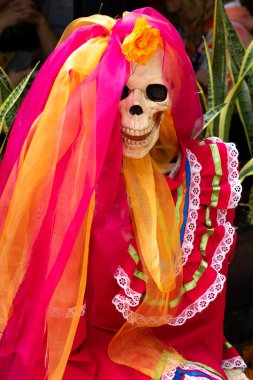Bir iskelet üzerinde çiçek olan kırmızı ve turuncu bir elbise giyer. İskelet peçe takıyor. Meksika Ölüler Günü geleneği, adaklar, yemek, renkler, dekorasyonlar ve papel picado, La Catrina.