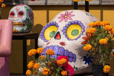 Portakal çiçekleriyle dolu bir vazonun yanındaki masaya büyük, boyalı bir kafatası yerleştirilir. Meksika Ölüler Günü geleneği, adaklar, yemek, renkler, dekorasyonlar ve papel picado, La Catrina.
