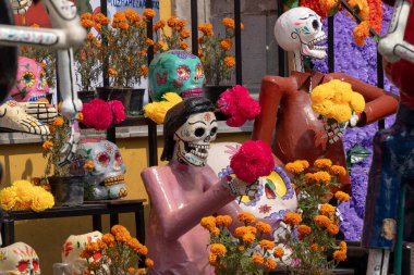 Bir grup iskelet çiçek tutuyor, bazıları maske takıyor. Sahne rengarenk. Meksika Ölüler Günü geleneği, adaklar, yemek, renkler, dekorasyonlar ve papel picado, La Catrina.