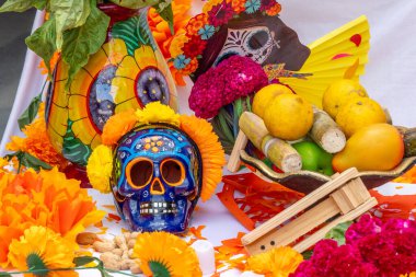 Kafatası, çiçekler ve meyveli bir masa. Kafatası çiçeklerle süslenmiş ve sarı bir yüze sahiptir. Meksika Ölüler Günü geleneği, adaklar, yemek, renkler, dekorasyonlar ve papel picado, La Catrina.