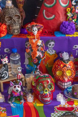 Kafatasları, iskeletleri ve kasvetli bir kadın dahil çeşitli cadılar bayramı süsleriyle kaplı bir masa. Meksika Ölüler Günü geleneği, adaklar, yemek, renkler, dekorasyonlar ve papel picado, La Catrina.