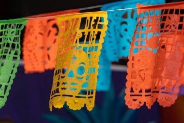 Üzerinde kafatası olan renkli bir pankart bir ipe asılı. Sancak kağıttan yapılmıştır. Meksika Ölüler Günü geleneği, adaklar, yemek, renkler, dekorasyonlar ve papel picado, La Catrina.