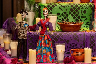 İçinde iki iskelet ve birkaç bardak olan bir masa. Masa bir mor ile süslenmiş. Meksika Ölüler Günü geleneği, adaklar, yemek, renkler, dekorasyonlar ve papel picado, La Catrina.