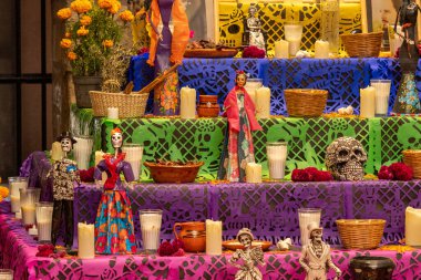 Renkli bir masa örtüsü ve üzerinde çeşitli dekorasyonlar olan bir yığın masa. Meksika Ölüler Günü geleneği, adaklar, yemek, renkler, dekorasyonlar ve papel picado, La Catrina.