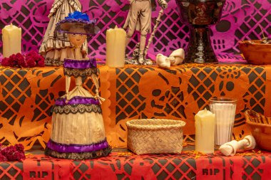 Mor bir elbise, şapka, kase, kupa ve mum giymiş bir bebek masası. Meksika Ölüler Günü geleneği, adaklar, yemek, renkler, dekorasyonlar ve papel picado, La Catrina.