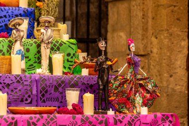 Çok renkli bir masa örtüsü ve bir yığın mumla dolu bir masa. Mumların üstünde. Meksika Ölüler Günü geleneği, adaklar, yemek, renkler, dekorasyonlar ve papel picado, La Catrina.