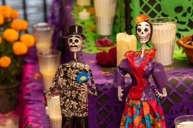 Siyah-beyaz ve mor giyinmiş iki iskelet diğer dekorasyonlarla birlikte bir masada. Meksika Ölüler Günü geleneği, adaklar, yemek, renkler, dekorasyonlar ve papel picado, La Catrina.
