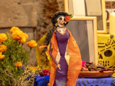 Turuncu ve mor giyinmiş bir kadın elinde çiçekler ve kafataslarıyla bir masanın önünde duruyor. Meksika Ölüler Günü geleneği, adaklar, yemek, renkler, dekorasyonlar ve papel picado, La Catrina.