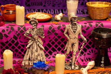 Beyaz ve siyah giyinmiş iki iskelet, mumlar ve diğer dekorasyonlarla dolu bir masanın üzerinde duruyor. Meksika Ölüler Günü geleneği, adaklar, yemek, renkler, dekorasyonlar ve papel picado, La Catrina.