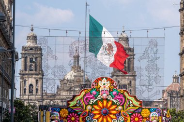 Kırmızı, beyaz ve yeşil renkli büyük bir bayrak büyük bir flo ile bir binanın önünde dalgalanıyor. Meksika Ölüler Günü geleneği, adaklar, yemek, renkler, dekorasyonlar ve papel picado, La Catrina.