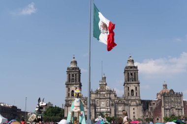 Büyük bir binanın önünde büyük bir bayrak dalgalanıyor. Bayrak kırmızı ve yeşil. Meksika Ölüler Günü geleneği, adaklar, yemek, renkler, dekorasyonlar ve papel picado, La Catrina.