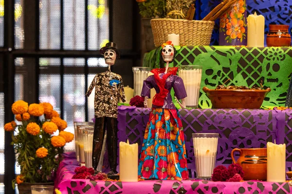 Bir erkek ve bir kadın kostüm giyer ve başka dekorasyonlarla dolu bir masada otururlar. Meksika Ölüler Günü geleneği, adaklar, yemek, renkler, dekorasyonlar ve papel picado, La Catrina.