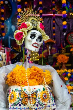 Turuncu çiçekler ve taçla süslenmiş beyaz bir elbise giymiş bir iskelet. Meksika Ölüler Günü geleneği, adaklar, yemek, renkler, dekorasyonlar ve papel picado, La Catrina.