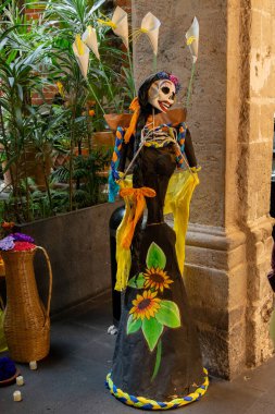 Elbisesinde sarı bir çiçek olan siyah ve sarı giyinmiş bir kadın. O bir iskelet. Meksika Ölüler Günü geleneği, adaklar, yemek, renkler, dekorasyonlar ve papel picado, La Catrina.