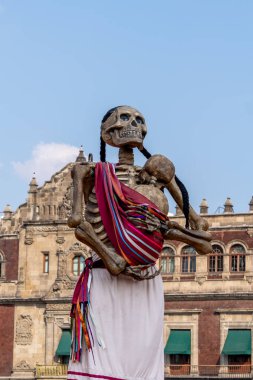 Bir iskelet kırmızı ve beyaz çizgili bir elbise giyer ve bir bebek tutar. Meksika Ölüler Günü geleneği, adaklar, yemek, renkler, dekorasyonlar ve papel picado, La Catrina.