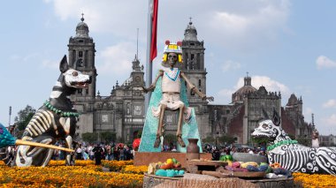 İskelet kostümü giymiş bir adam büyük bir binanın önünde bir kaidede oturur. Meksika Ölüler Günü geleneği, adaklar, yemek, renkler, dekorasyonlar ve papel picado, La Catrina.