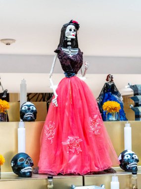 Bir iskelet pembe bir elbise giyiyor ve üzerinde birkaç kafatası olan bir masanın önünde duruyor. Meksika Ölüler Günü geleneği, adaklar, yemek, renkler, dekorasyonlar ve papel picado, La Catrina.