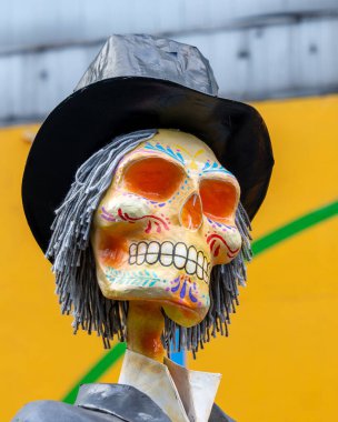 Kafasında şapka olan bir iskelet. Şapka siyahtır ve iskelet parlak renklerle boyanmıştır. Meksika Ölüler Günü geleneği, adaklar, yemek, renkler, dekorasyonlar ve papel picado, La Catrina.