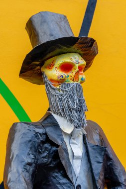 Yüzünde kafatası olan takım elbiseli ve şapkalı bir adam. Kafatası sarı ve kırmızıdır. Meksika Ölüler Günü geleneği, adaklar, yemek, renkler, dekorasyonlar ve papel picado, La Catrina.
