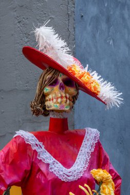 Bir iskelet kırmızı bir elbise ve beyaz bir şapka giyiyor. Elbise dantelle süslenmiş. Meksika Ölüler Günü geleneği, adaklar, yemek, renkler, dekorasyonlar ve papel picado, La Catrina.