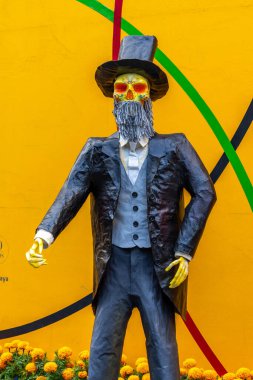 Yüzünde kafatası olan takım elbiseli ve şapkalı bir adam sarı bir duvarın önünde duruyor. Meksika Ölüler Günü geleneği, adaklar, yemek, renkler, dekorasyonlar ve papel picado, La Catrina.
