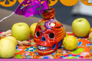 Bir kafatası elma ve portakallarla dolu bir masada oturuyor. Kafatası parlak renklerle boyanmıştır. Meksika Ölüler Günü geleneği, adaklar, yemek, renkler, dekorasyonlar ve papel picado, La Catrina.
