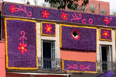 Çiçekler ve sarmaşıklarla kaplı bir bina. Çiçekler mor ve kırmızıdır. Meksika Ölüler Günü geleneği, adaklar, yemek, renkler, dekorasyonlar ve papel picado, La Catrina.