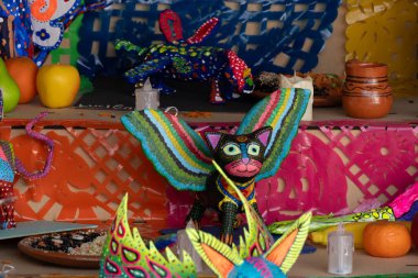 Masanın üzerinde renkli bir kedi heykelciği ve diğer süslemeler duruyor. Kedi bir sopa tutuyor. Meksika Ölüler Günü geleneği, adaklar, yemek, renkler, dekorasyonlar ve papel picado, La Catrina.