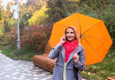 Güzel sonbahar park telefonunu kullanarak şemsiye ile genç kadın.