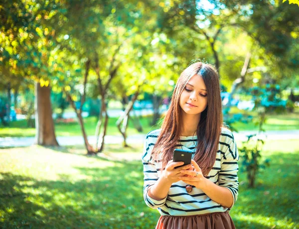 Happy beautiful young Caucasian girl with smart phone outdoors on sunny summer day tex-ting and smiling — Stock Photo, Image
