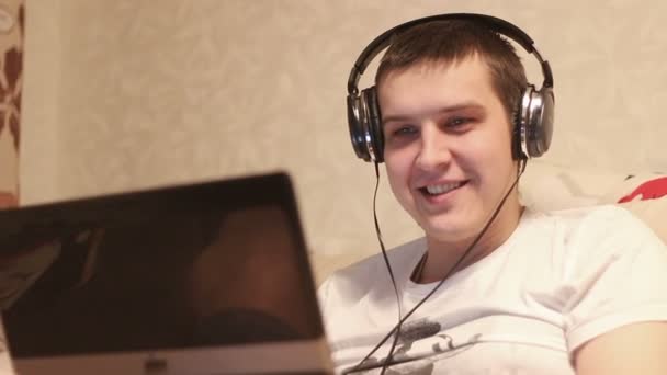 Νεαρός άνδρας κάθεται στον καναπέ με ένα φορητό υπολογιστή και να ακούσετε μουσική στα ακουστικά — Αρχείο Βίντεο