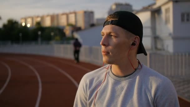 La cámara lenta del joven usa auriculares en el campo al atardecer en la pista del estadio — Vídeos de Stock