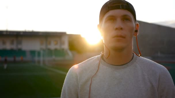 Slowmotion of young man wears headphones on field at sunset on stadium track — Stock Video