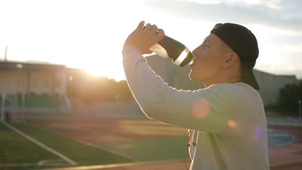 L'athlète fatigué avec une casquette de baseball boit de l'eau de la bouteille sur la piste du stade — Video