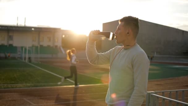 Уставший спортсмен в носовой кепке пьет воду из бутылки на стадионной дорожке — стоковое видео