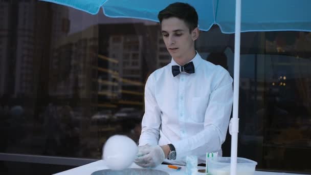 Young guy making cotton candy on a special machine, it wears bow tie, behind him balloons — Stock Video