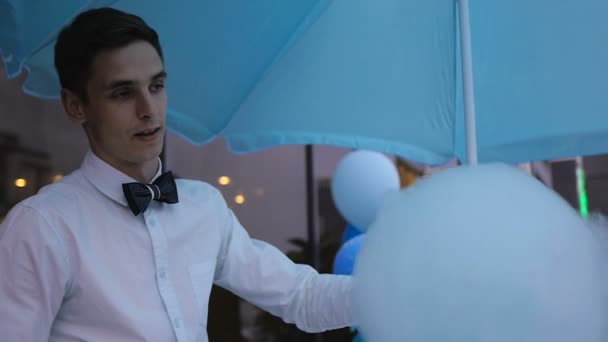 Young guy making cotton candy on a special machine, it wears bow tie, behind him balloons — Stock Video