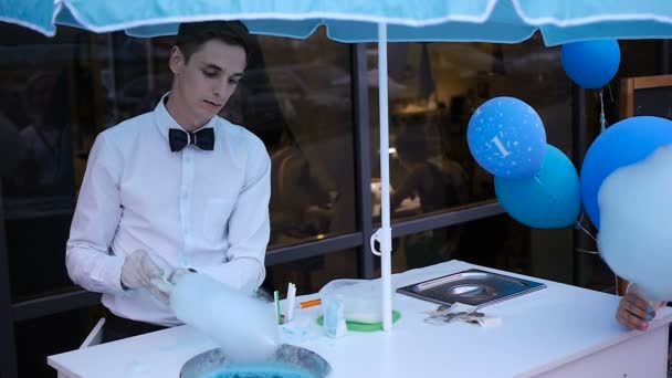 Chico joven haciendo algodón de azúcar en una máquina especial, lleva corbata de lazo, detrás de él globos — Vídeos de Stock