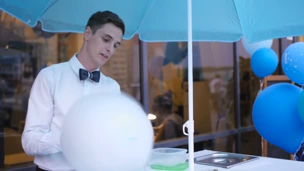 Junger Kerl, der Zuckerwatte an einer speziellen Maschine herstellt, trägt Fliege, hinter ihm Luftballons — Stockvideo