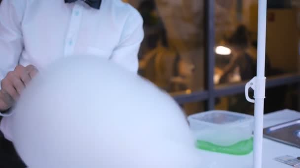 Chico joven haciendo algodón de azúcar en una máquina especial, lleva corbata de lazo, detrás de él globos — Vídeos de Stock