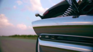 Sakallı orta yaşlı motorcu gün batımında motosikletin yanında oturur.