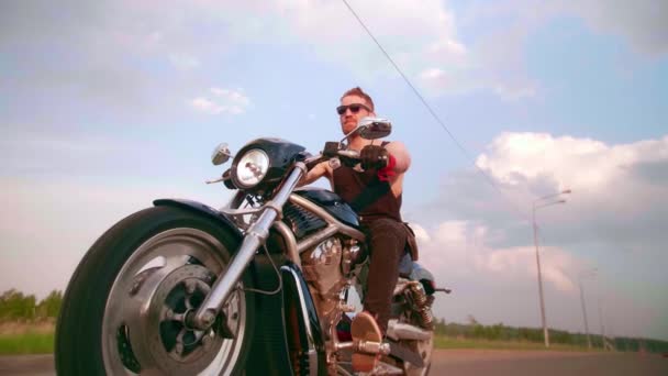 Elegante motociclista con tatuajes monta una motocicleta en una carretera de campo al atardecer — Vídeo de stock